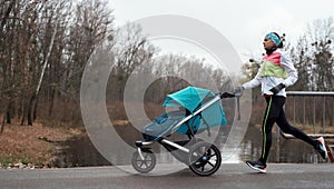 Side view of sporty young woman mom running with a stroller along an empty park on a cold autumn day