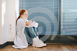 Side view of stressed upset female doctor feeling worried about professional malpractice sitting on floor near window in