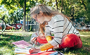 Side view of student female in casual outfit sitting on the green grass at the college campus, and studying with lots of books