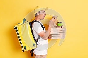 Side view of surprised male courier wearing cap and holding thermo bag holding pizza boxes from restaurant and using smartphone,