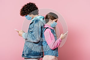 Side view of teenagers in denim jackets and medical masks using smartphones on pink background.