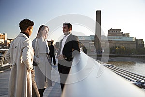 Side view of three millennial business colleagues stand talking on Millennium Bridge, London, selective focus