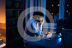 Side view of tired young business man working on computer in dark home office at night