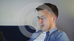 Side view a tired young man is lying on the couch and working on a laptop, typing on the keyboard, remote work from home