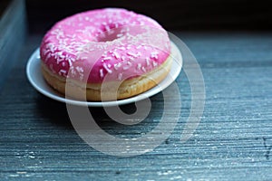 side view to bright pink donut on white round plate and aged wooden grey background. Isolated object close up
