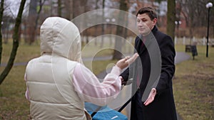 Side view troubled man arguing with angry woman standing with baby stroller on park alley. Dissatisfied Caucasian wife