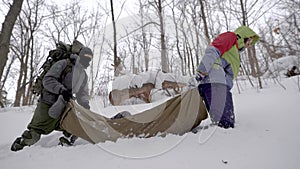 Side view of two man with trekking equipment dragging injured woman on a stretcher in the forest in winter.