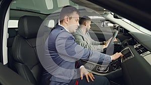 Side view of two men customer and salesman sitting in beautiful car on front seats and talking touching car parts