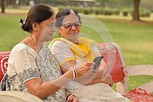 Side view of two Senior Indian woman working on mobile phone/tablet, experimenting with technology on a red park bench in an