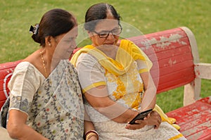 Side view of two Senior Indian woman working on mobile phone/tablet, experimenting with technology on a red park bench in an