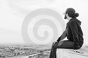 Side view unidentified young man in casual clothes and glasses sits on the high rock and looks at beautiful desert