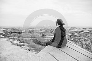 Side view unidentified young man in casual clothes and glasses sits on the high rock and looks at beautiful desert