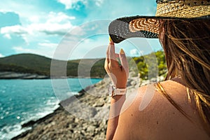 Side view of an unrecognizable brunette girl, holding onto her hay hat in the summer. Standing near the shore of the adriatic sea