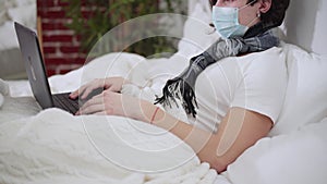 Side view of unrecognizable ill Caucasian man in coronavirus face mask lying on bed couching and talking using video