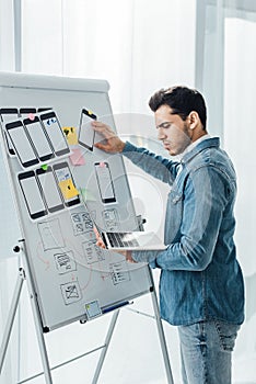 Side view of ux designer using laptop near layouts of mobile frameworks on whiteboard