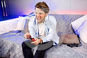 Side view of venturous millennial male sit on neon sofa at home, having fun playing video games with red game pad photo