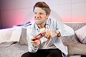 Side view of venturous millennial male sit on neon sofa at home, having fun playing video games with red game pad photo
