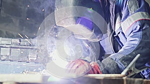 Side view welder in welding mask welds two metal parts. Workman in coveralls is working indoors