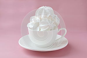 Side view on white meringues in a white teacup on a white plate on a pink background