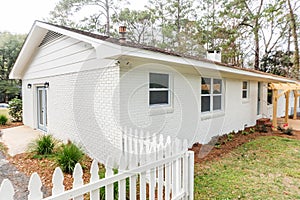 Side view of a white ranch house with a short white picket fence surrounding a backyard and paved driveway