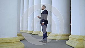 Side view wide shot of young African American man in suit walking between white columns holding smartphone and looking