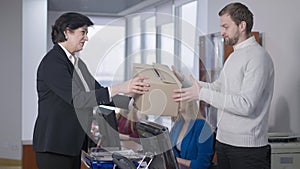 Side view of woman giving fired man cardboard box for packing. Serious Caucasian female CEO firing unprofessional