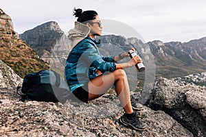 Side view of woman hiker in sports clothes enjoying the view. Female in sunglasses holding a thermos sitting on a rock and