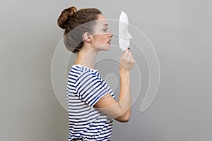 Side view of woman holding and looking at white mask with attentive look, trying to hide personality