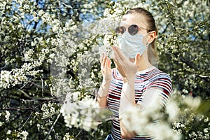 Side view of woman in medical mask and sunglasses on background of blossoming apple tree on summer day.