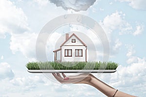 Side view of woman`s hand holding tablet with green grass and little detached house on screen, with rain cloud above