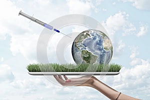 Side view of woman`s hand holding tablet with green grass on screen and little Earth globe lying on it with syringe in