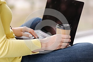 Side view on woman workin on laptop and hold cup of coffee. Female in yellow sweeter sitting on window sill