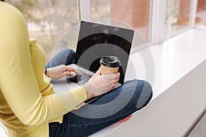 Side view on woman workin on laptop and hold cup of coffee. Female in yellow sweeter sitting on window sill