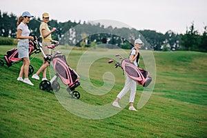 side view of women in polos and caps with golf gear walking on green lawn photo