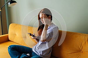 Side view of worried young woman sitting on sofa, received message with bad news, eyes closed and touching head.