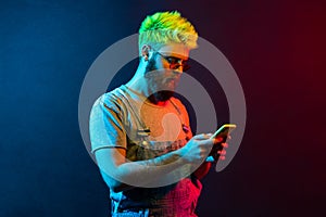 Side view of young adult bearded hipster man wearing denim overalls, holding smart phone in hands, looking at display with serious
