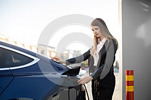 Side view of young adult woman holding in hand power cable supply plugged in electric car, charging vehicle battery at