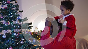 Side view young African American man decorating Christmas tree at home talking with toddler boy on shoulders. Curios