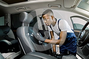 Side view of young African man, car service worker, wearing overalls and protective gloves, cleaning car front seat