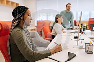 Side view of young African woman writing down notes and using computer sitting at work in big modern corporate office