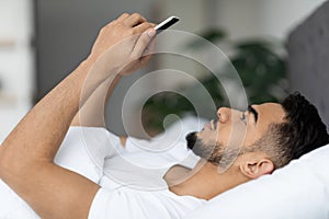 Side View Of Young Arab Man Using Smartphone While Lying In Bed