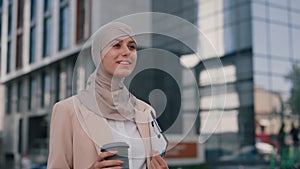 Side view of the young arabian elegant business woman wearing brown hijab holding coffee and notebook walking on the