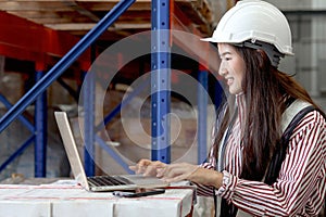Side view of young Asian beautiful woman wearing safety vest and helmet, typing on a laptop computer at cargo logistics warehouse