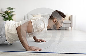 Side view of young athletic Arab man doing push-ups on sports mat at home, copy space