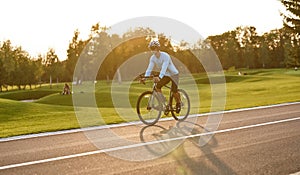 Side view of a young athletic man in sportswear cycling in city park at sunset, riding mountain road bike