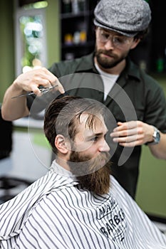 Side view of young bearded man getting beard haircut at hairdresser while sitting in chair at barbershop
