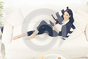 Side view of Young beautiful Asian woman look at the camera sitting on white sofa using tablet smartphone explore website. reading