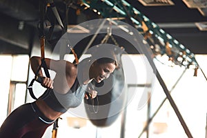 Side view of young beautiful fitness woman in sport wear doing TRX workout at industrial gym