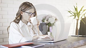 Side view of a young beautiful woman wearing glasses typing at her laptop in office. Medium shot.