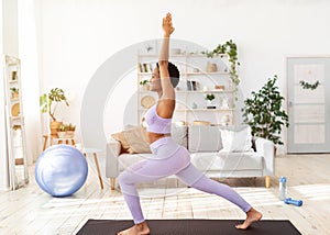 Side view of young black woman practicing yoga at home, standing in warrior pose, exercising her strength